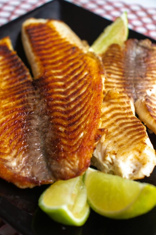 Delicious grilled tilapia fillets served with fresh lime wedges on a dark plate.